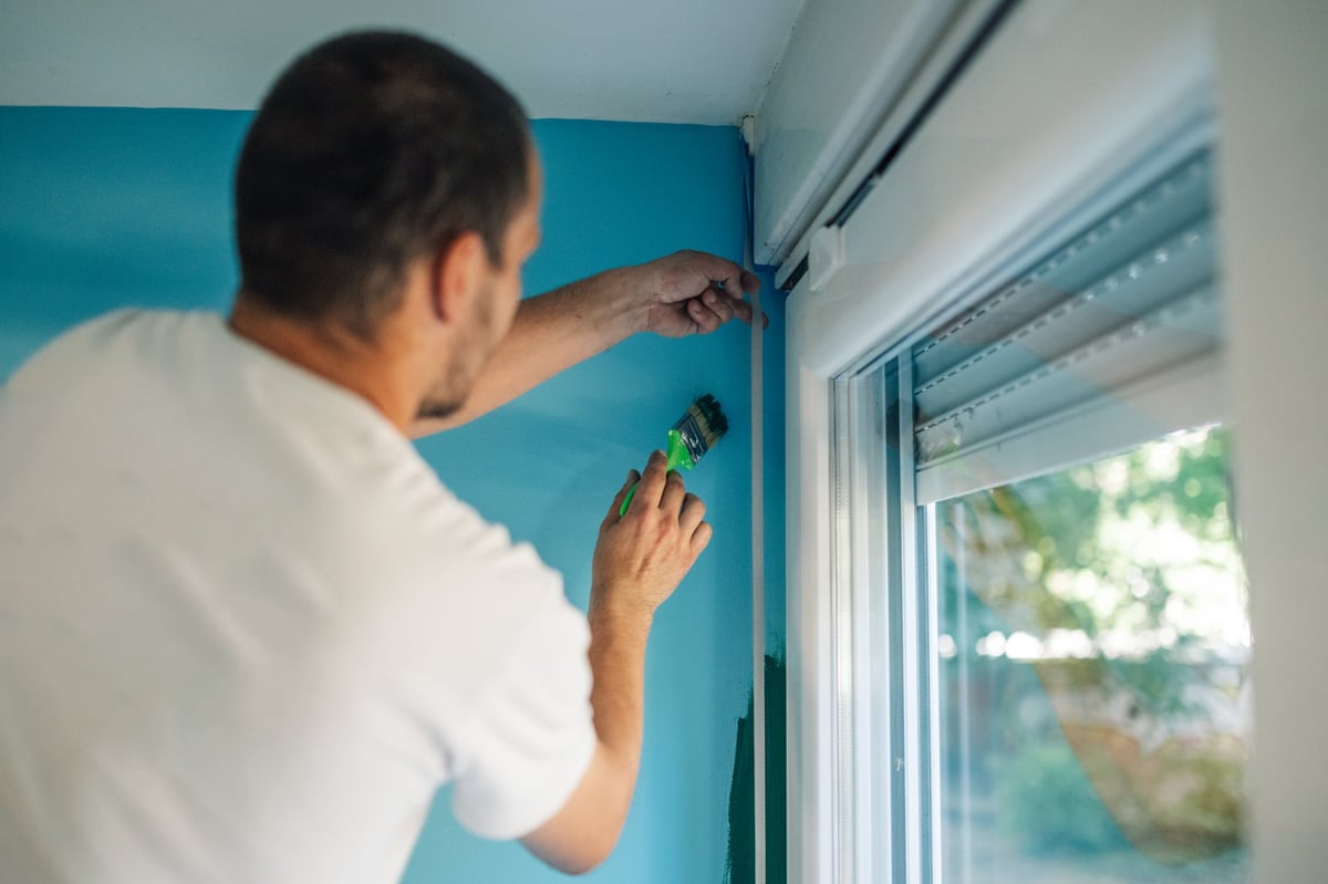 Painter applying masking tape around a window while painting a wall blue